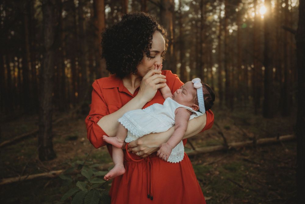 A woman in a red dress cradles a baby outdoors in a forest setting.