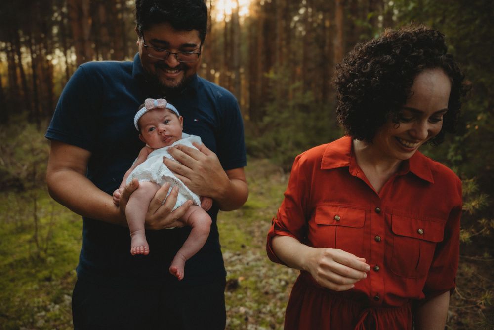 A couple holds their newborn baby while standing in a sunlit forest clearing.