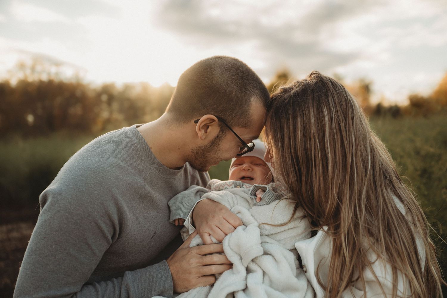 Family shares tender moment with their newborn baby in sunset-lit field.