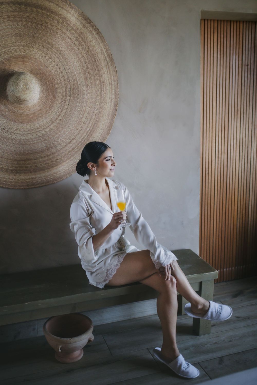 A person in a white robe sits on a bench holding an orange drink, with a large woven disc art piece on the wall.