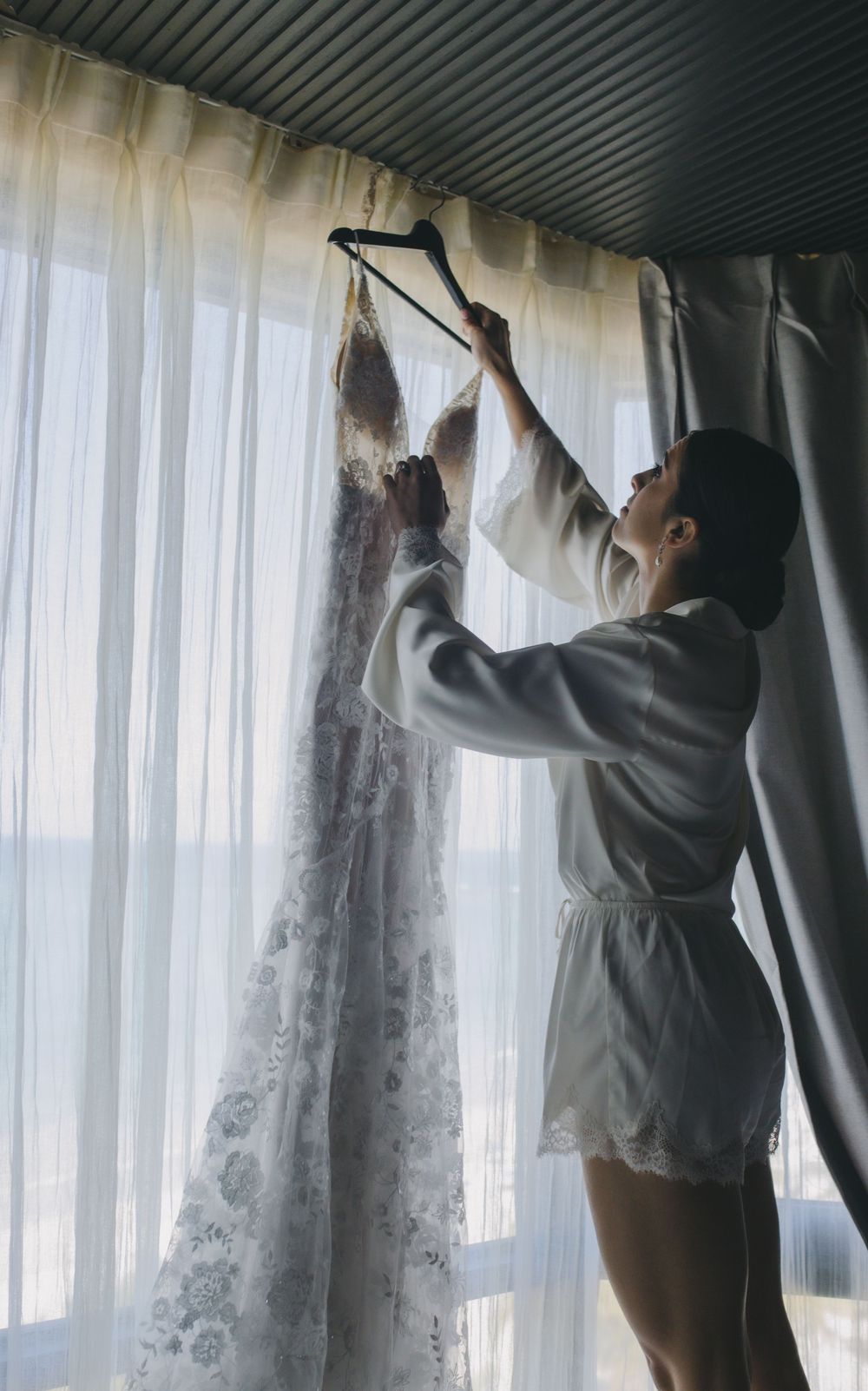 A person in a robe stands near a window hanging a delicate white lace wedding dress on a clothing hanger.