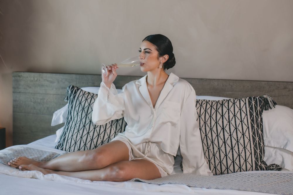 Person relaxing on bed in white bathrobe while sitting against pillows and sipping from glass.