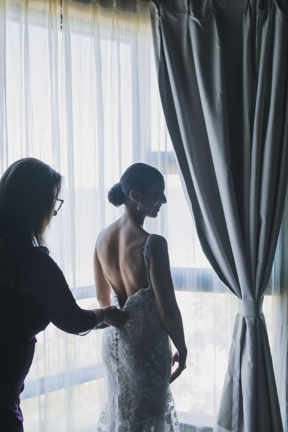 A silhouetted figure helps adjust a lace wedding dress near a window with sheer curtains and draped fabric.