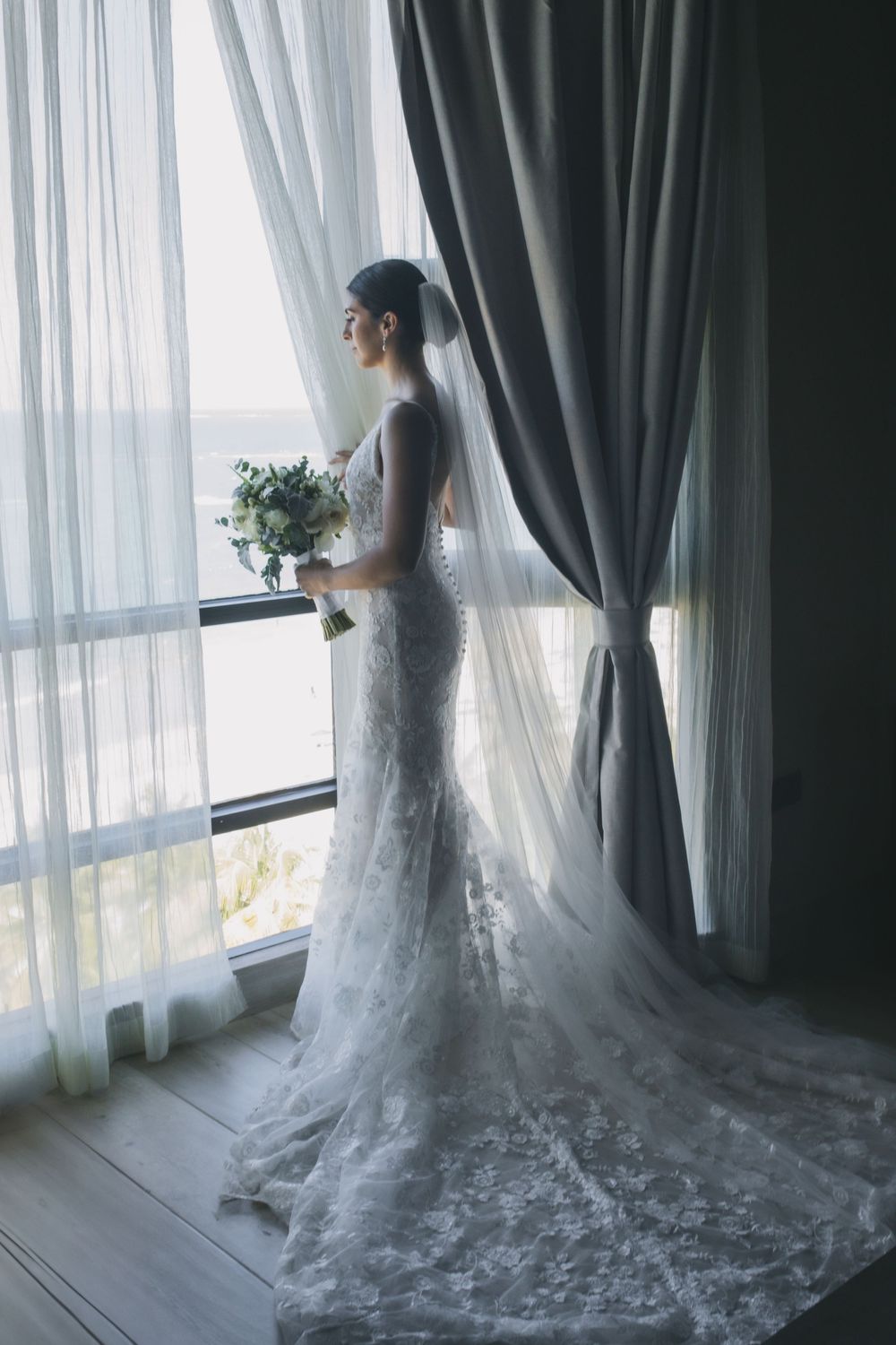 A bride in an elegant lace wedding gown with long train stands by window holding a white floral bouquet.