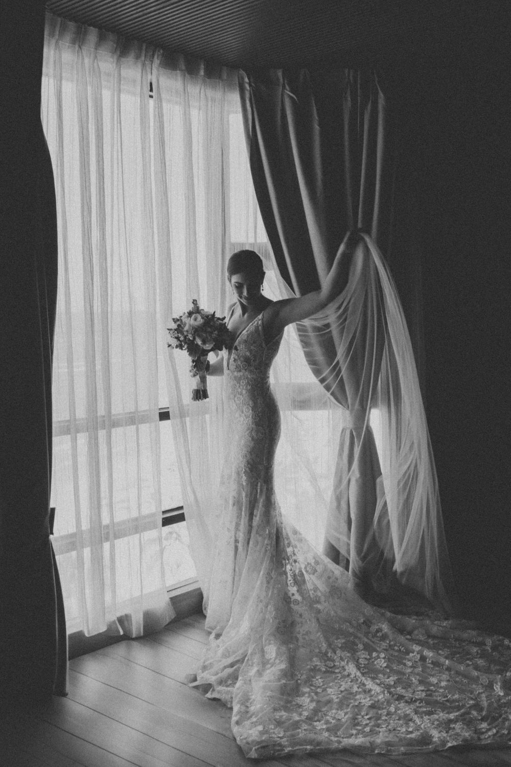 Silhouette of a bride holding a bouquet stands against a window with flowing curtains and long lace train in black and white.