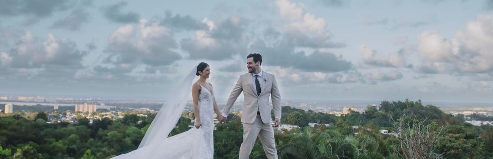 A couple on their wedding day stands at an overlook with a scenic cityscape view and cloudy sky in the background.