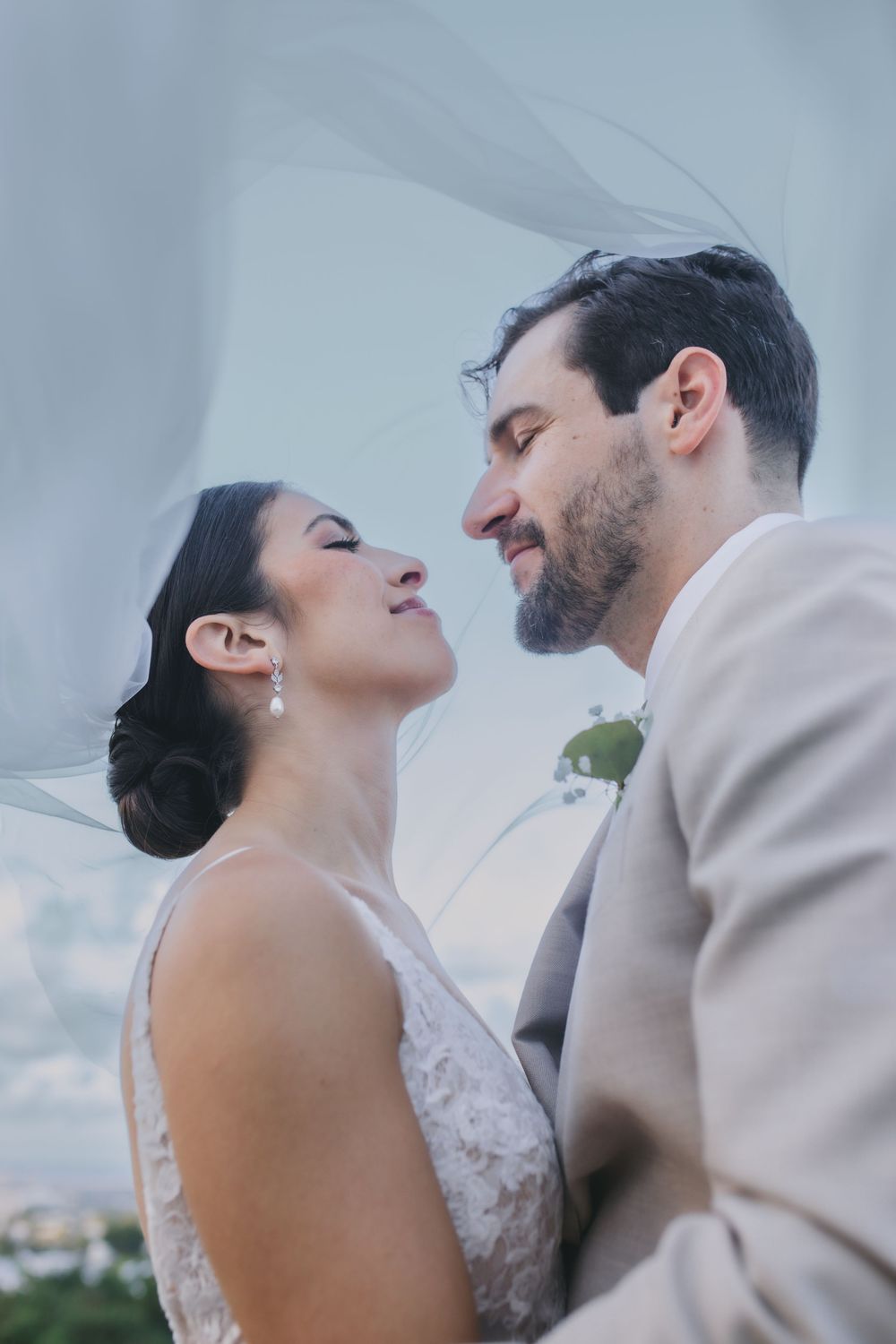A newly married couple in wedding attire share an intimate moment under a cloudy sky during their wedding photoshoot.