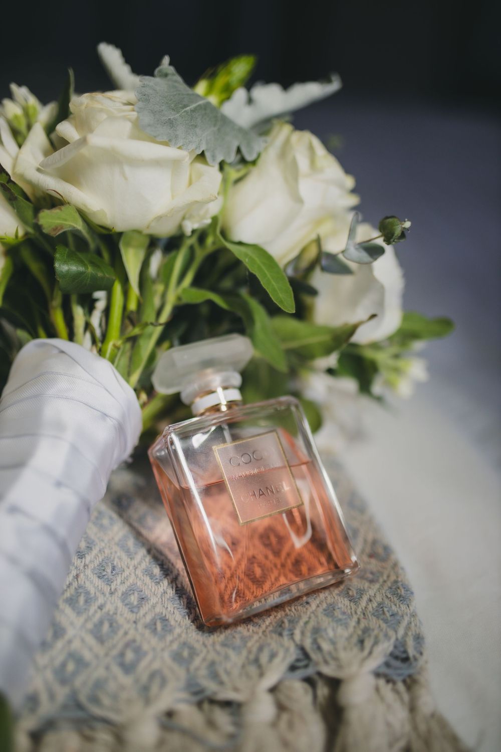 Elegant white roses in a bouquet next to a light pink perfume bottle on patterned fabric.