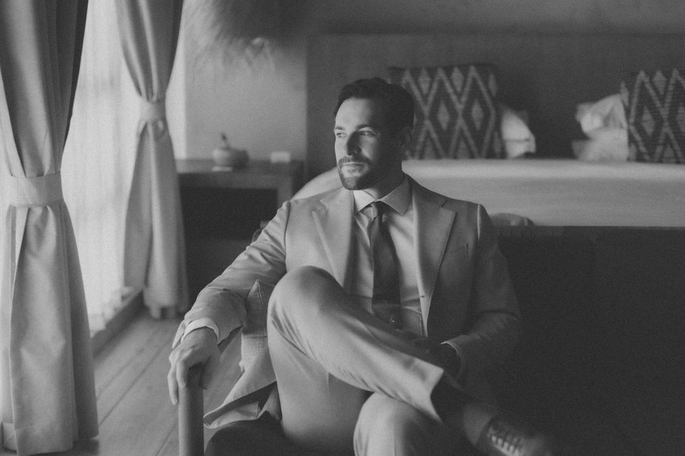 A person in a light suit sits comfortably in an armchair by a window in a monochrome photo.