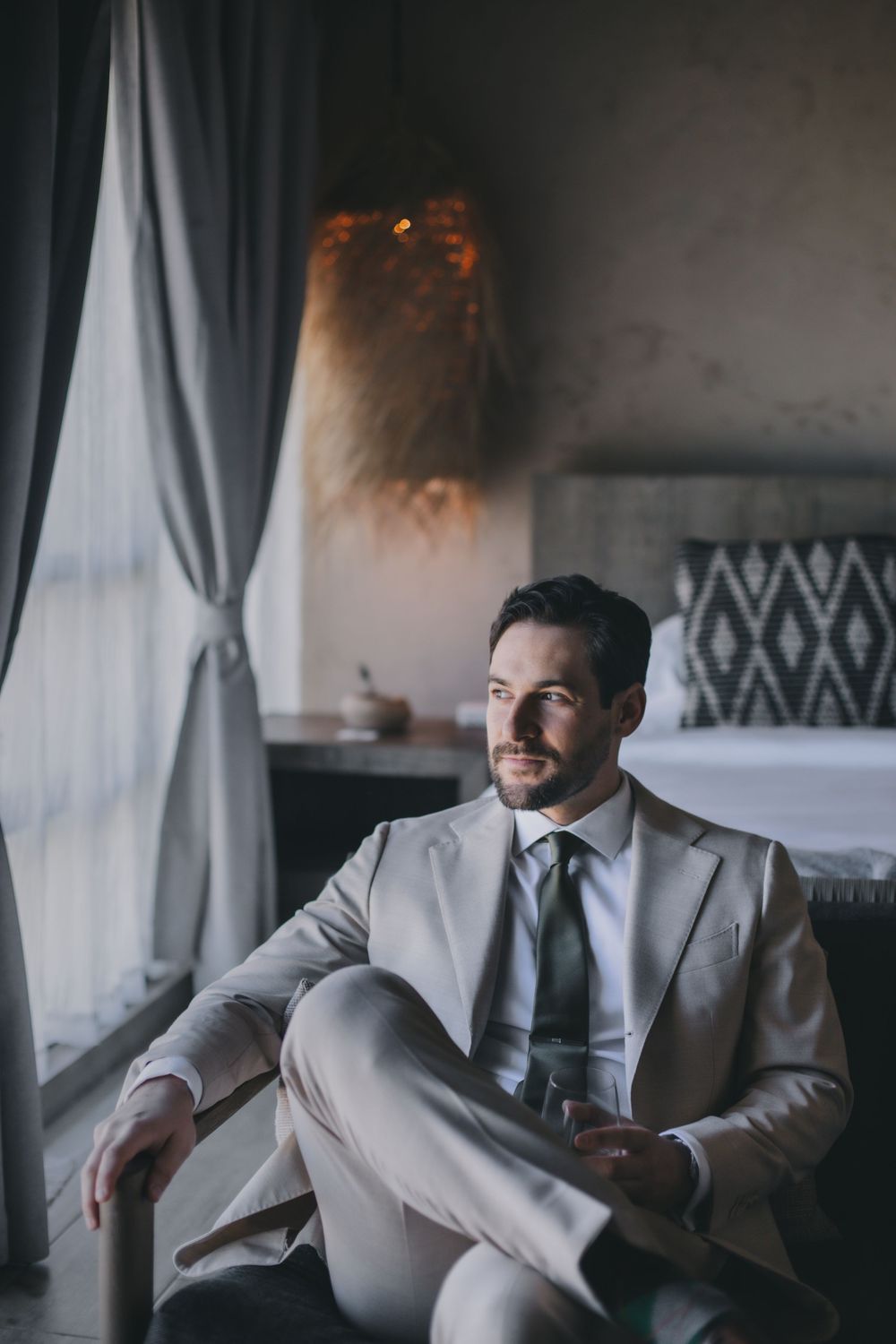 Professional in a light gray suit sits relaxed by a window in a moody, elegantly decorated room.