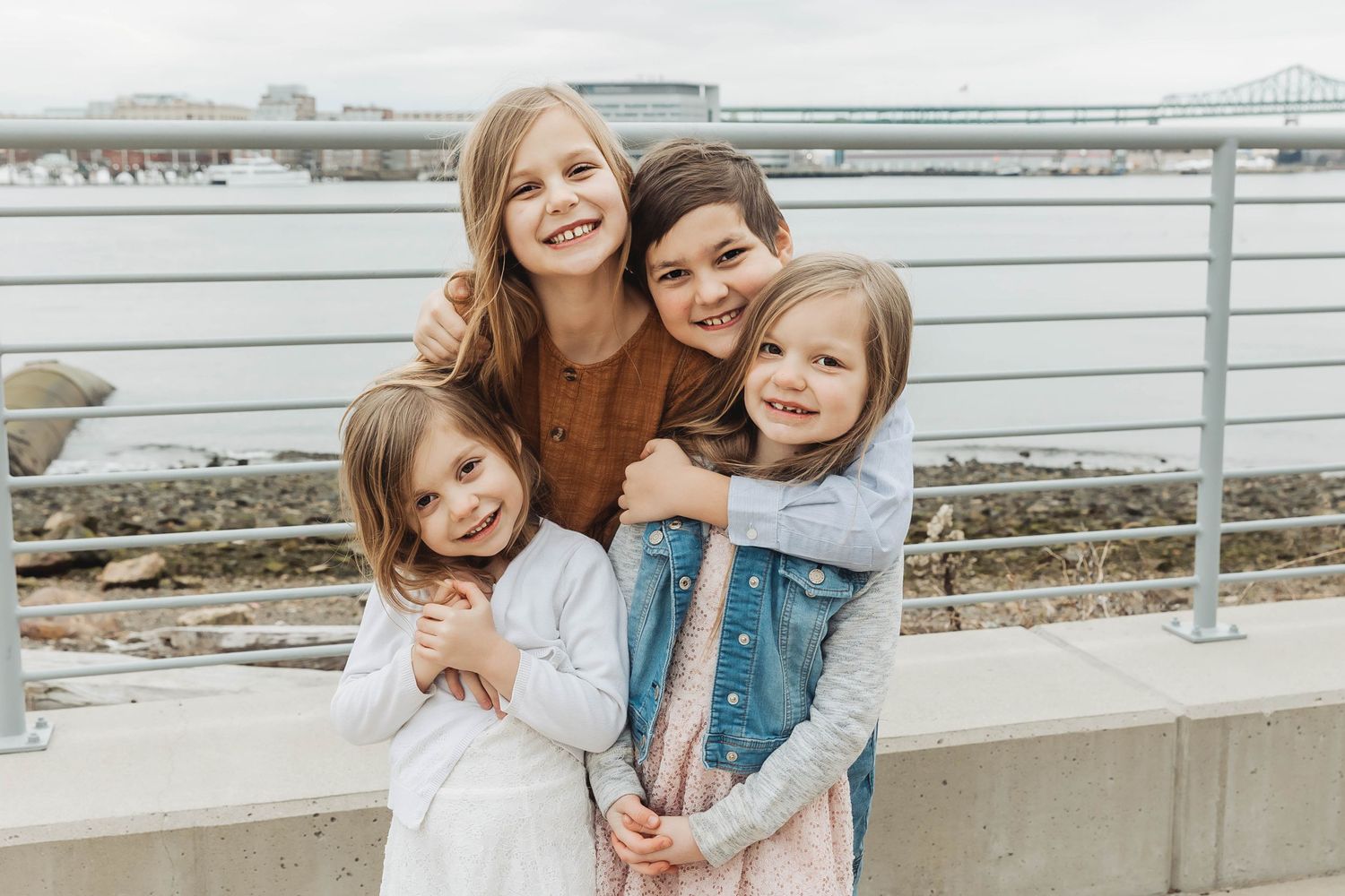Four children embrace and smile while standing against a waterfront railing on an overcast day.