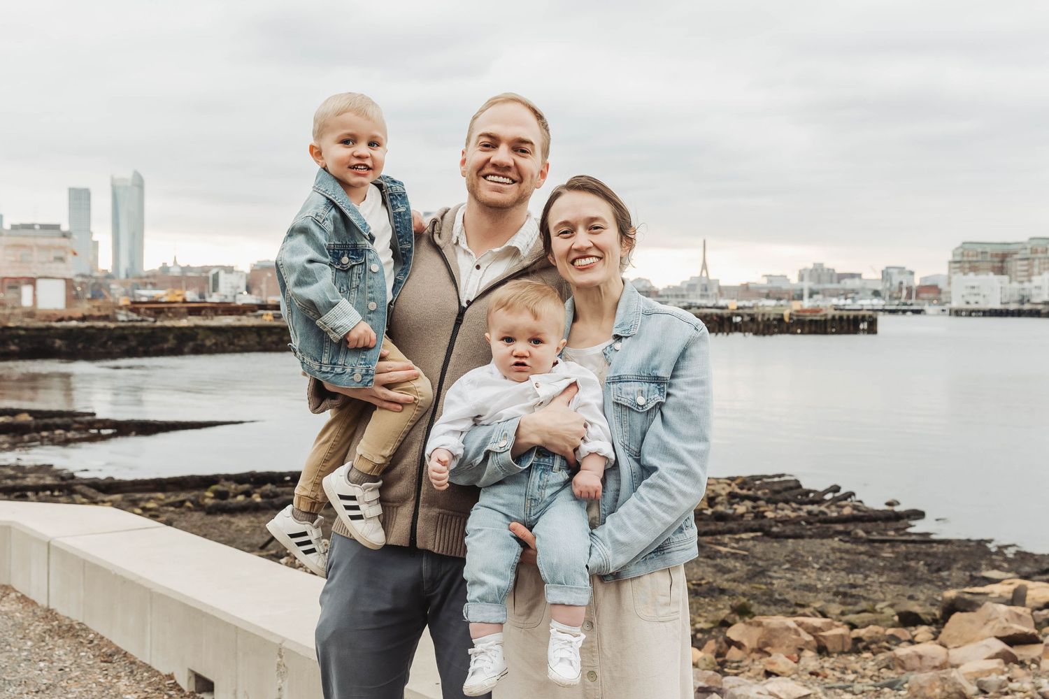 A family of four poses together near the water with a city skyline behind them.
