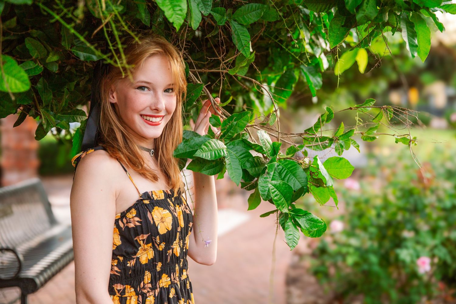 A person in a floral dress poses outdoors under green foliage in warm sunlight.