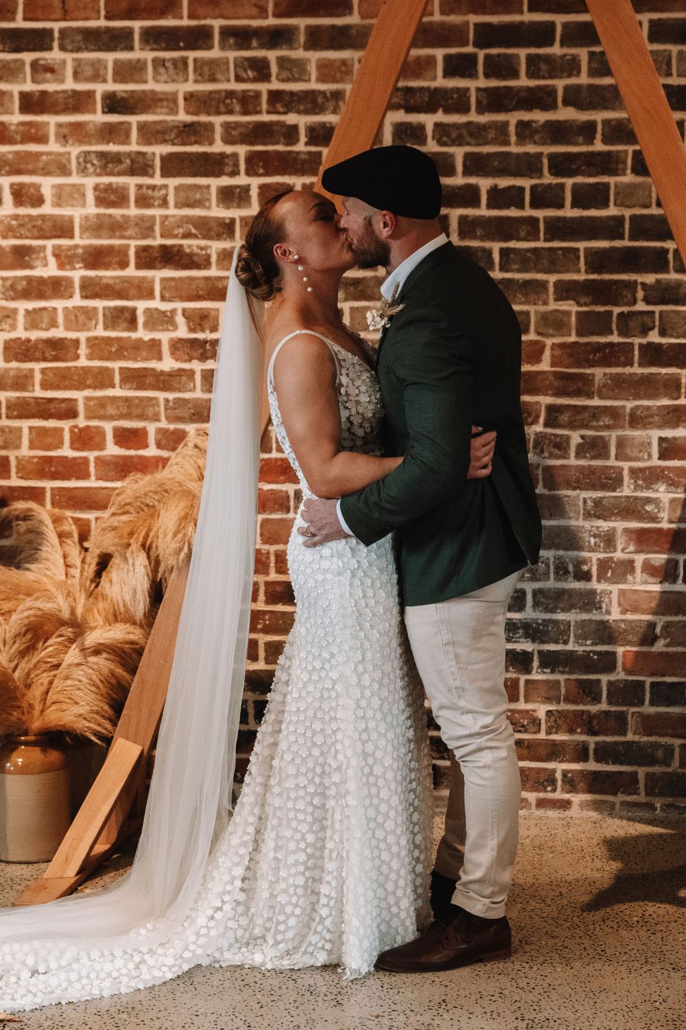 A romantic wedding moment between a couple in front of a brick wall with pampas grass decor.