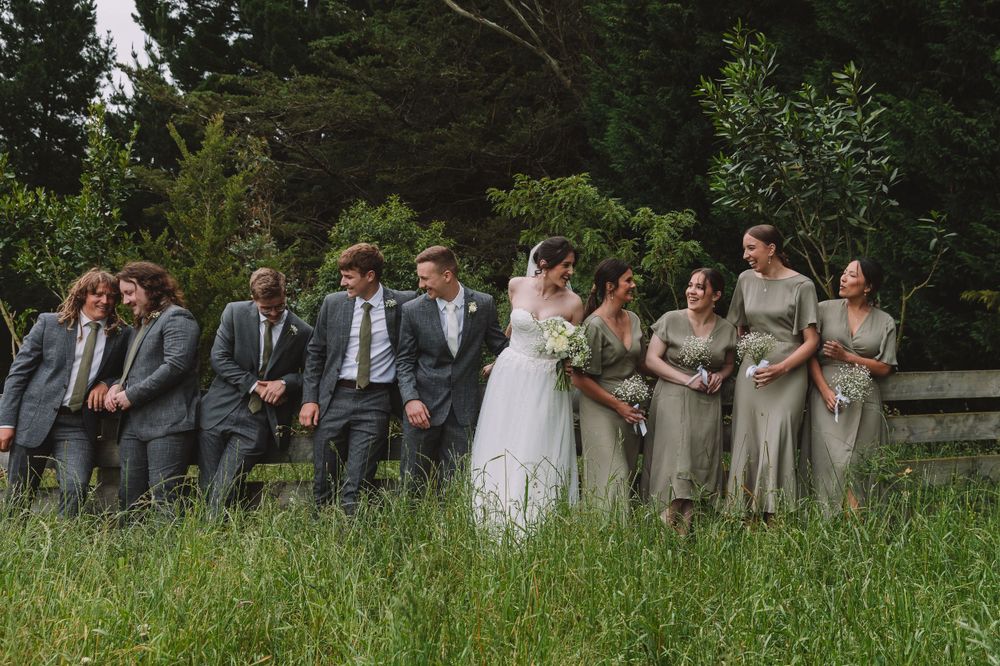 A wedding party poses together in a rustic outdoor setting with bridesmaids in sage dresses and groomsmen in gray suits.