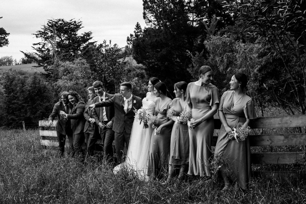 Black and white photo of a wedding party standing together in a grassy meadow by a fence.