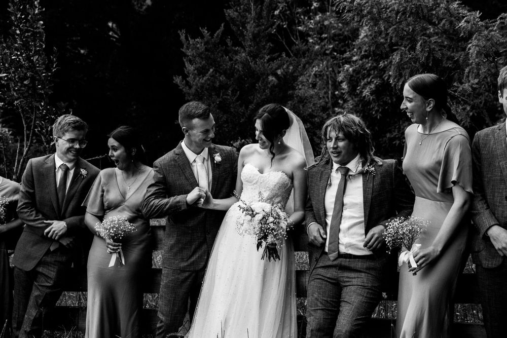 Black and white candid photo captures wedding party sharing laughs together outdoors.