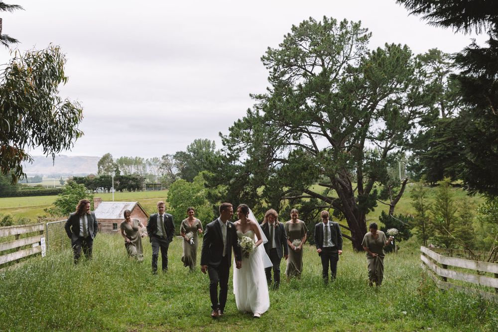 Wedding party walks together through a scenic meadow with rolling hills and trees in the distance.