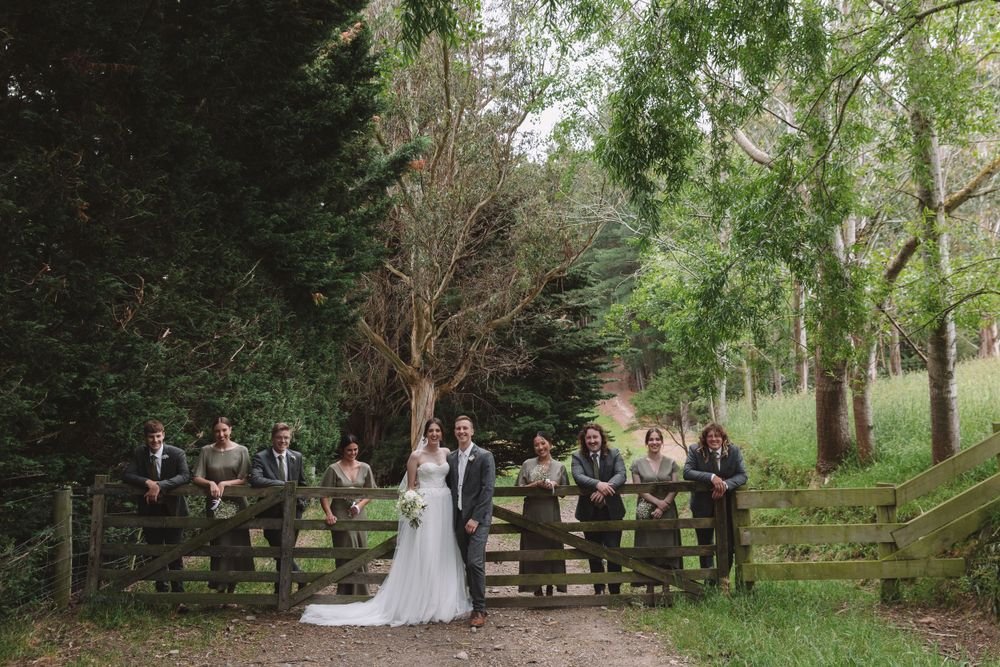 Wedding party poses against rustic wooden fence in a serene forest setting.