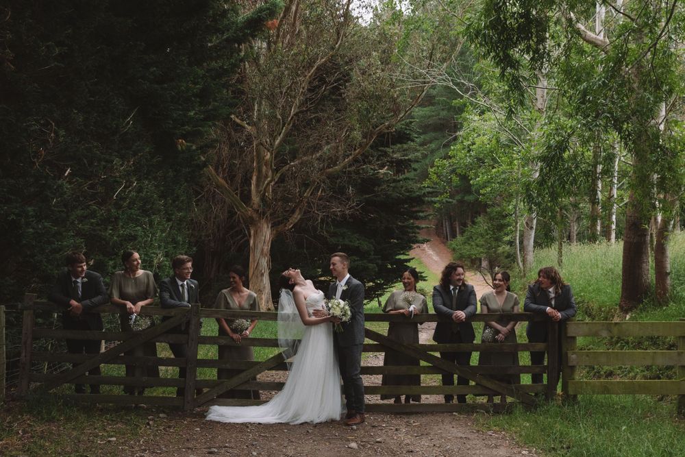 Wedding party gathers by a rustic wooden fence in a moody forest setting.