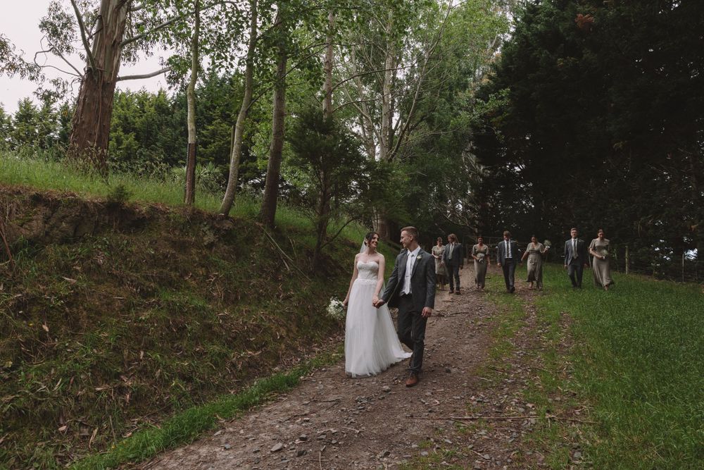 Wedding procession walks along a scenic wooded trail surrounded by trees and greenery.