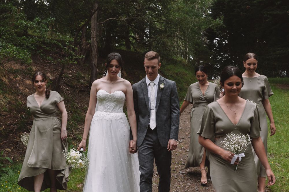 Wedding party walks together on a forest path wearing elegant attire and carrying bouquets.