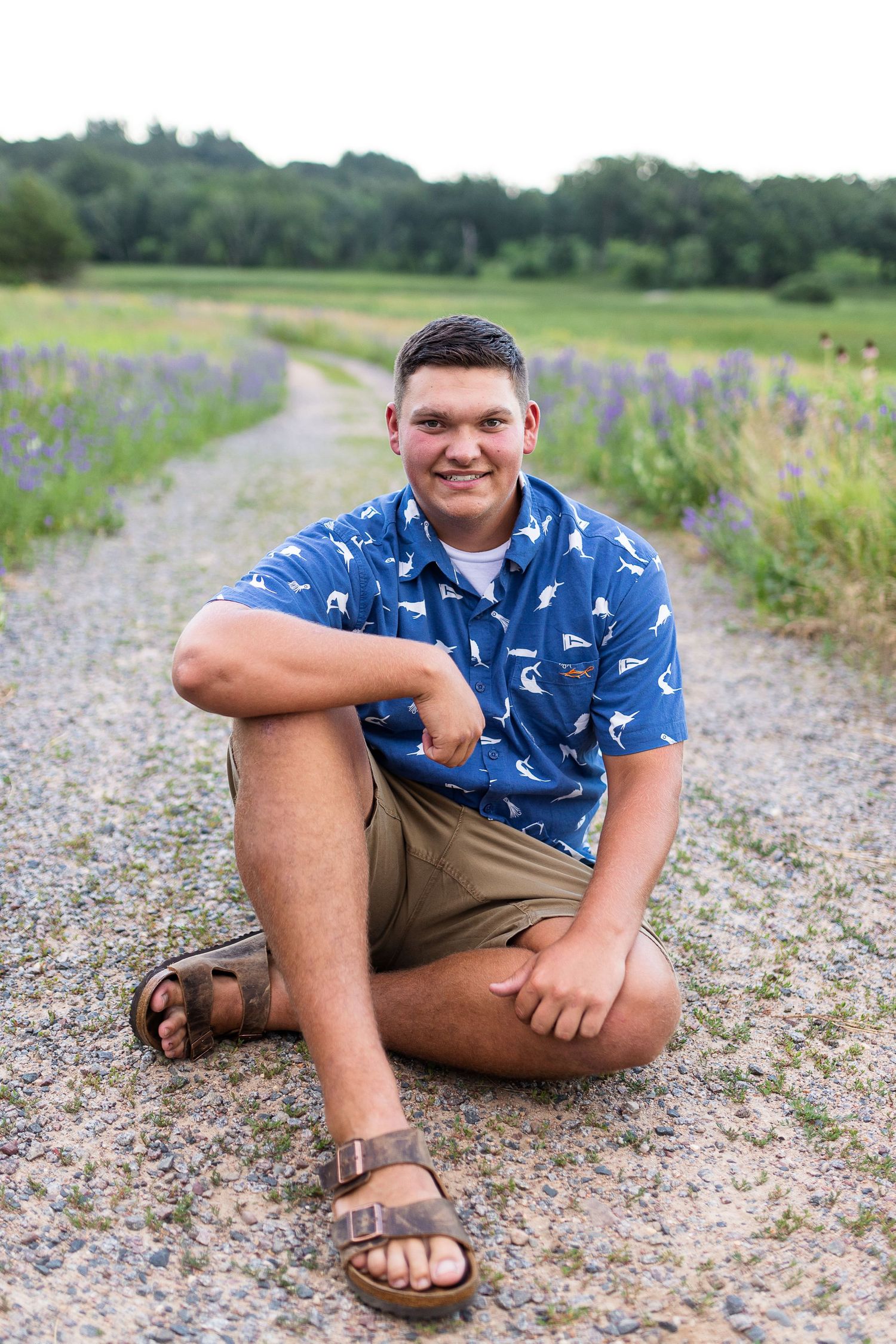 Male Senior sitting on the ground in a sword fish shirt. Path of purple flowers behind him. Captured by Shania Hirdler