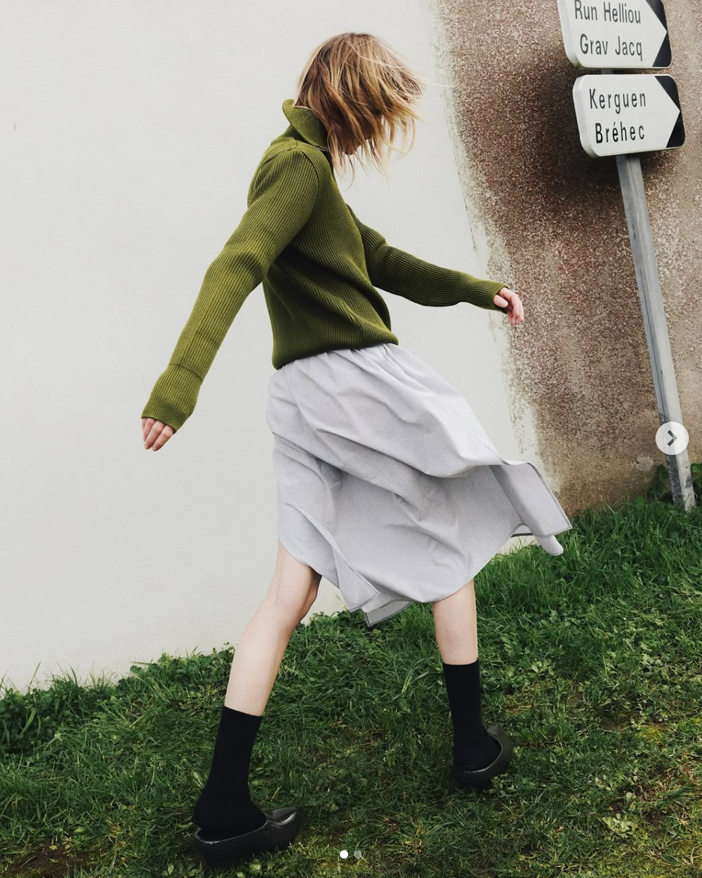 Figure in olive sweater and flowing gray skirt dances near street sign.