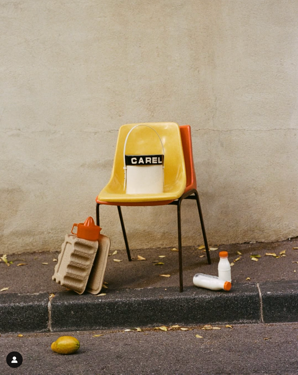 Yellow vintage chair labeled 'Carel' sits on sidewalk with orange accents and scattered objects.