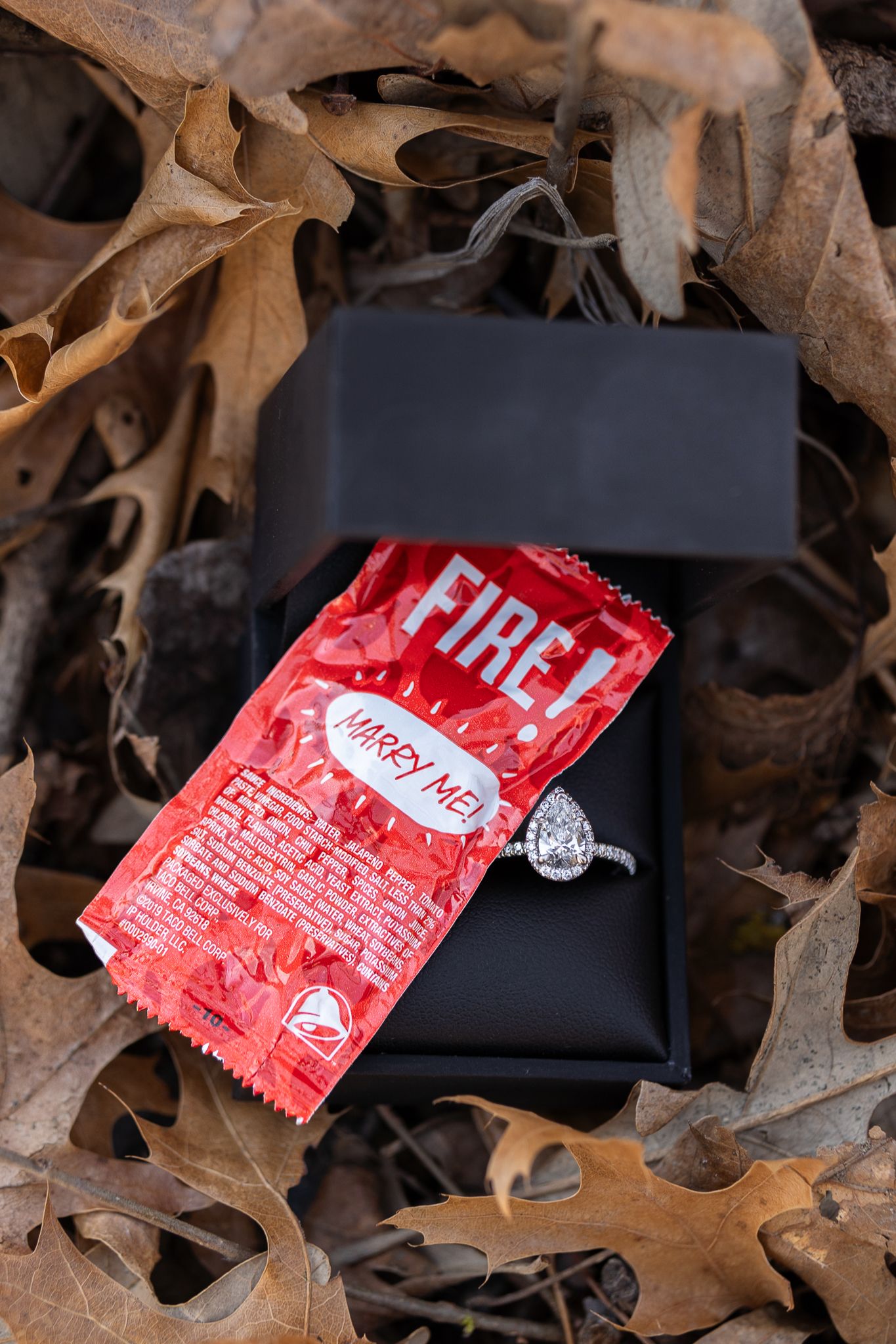 Diamond ring and Taco Bell Fire sauce packet displayed on black box among fallen leaves.