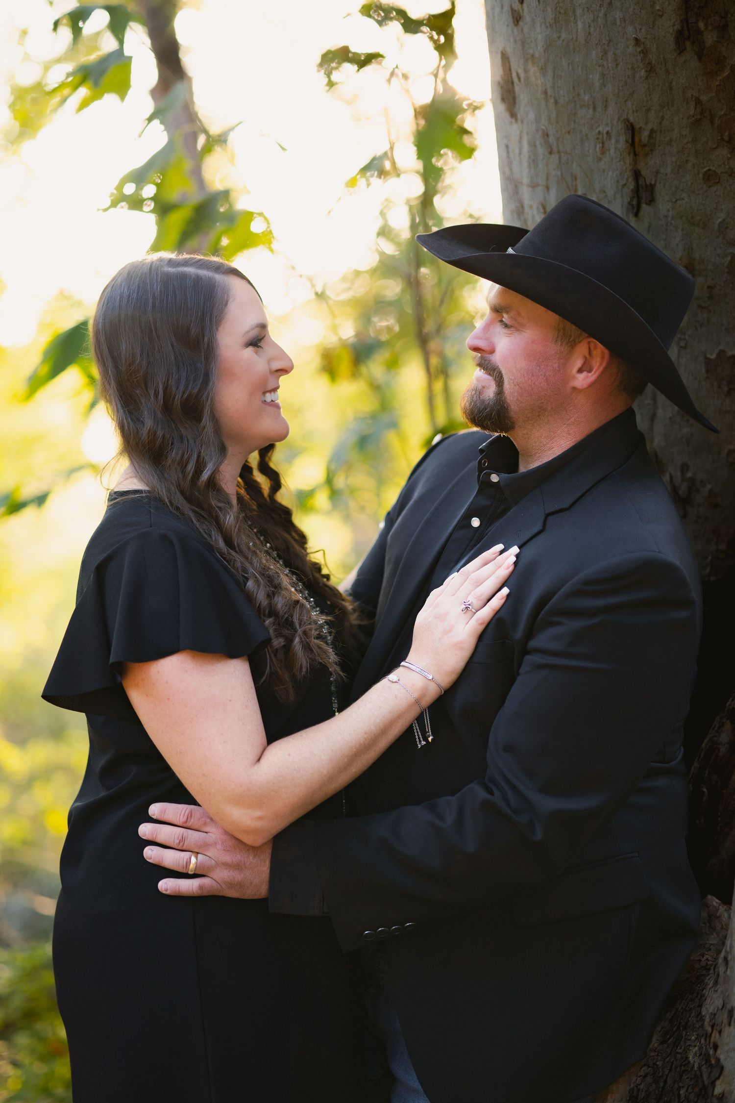 A romantic couple in black outfits pose together against a sunlit natural backdrop.