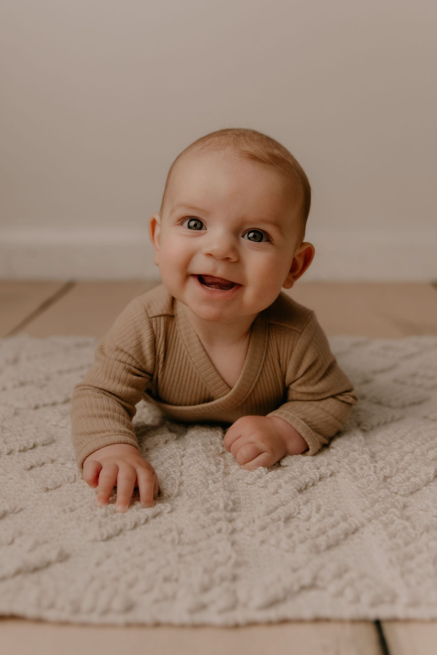 A baby in a tan sweater lies on a textured cream-colored rug while smiling joyfully.