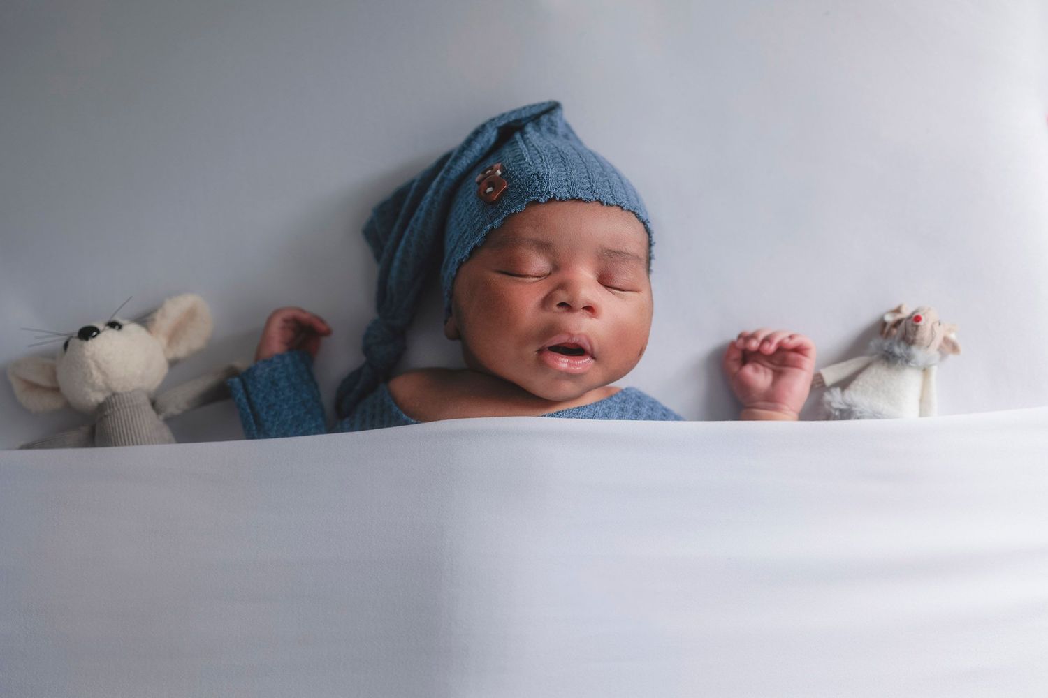 Sleeping newborn in blue cap rests between small stuffed animals on white. Hampshire newborn photographer, Gosport Newborn baby photography