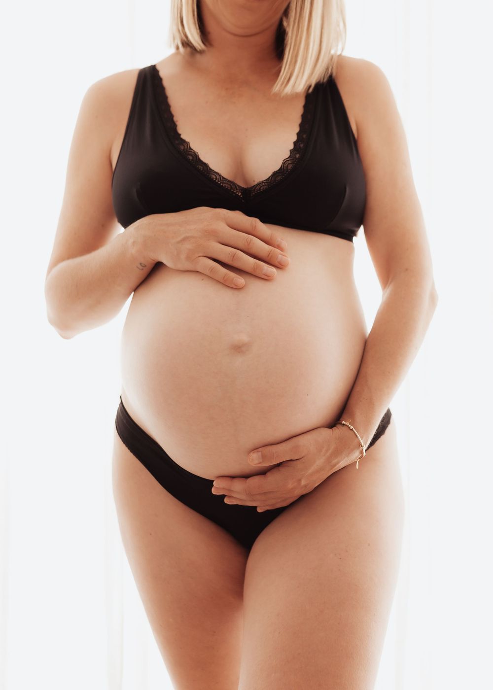 Pregnant woman in black lingerie poses against white background while holding her belly in a maternity photo.