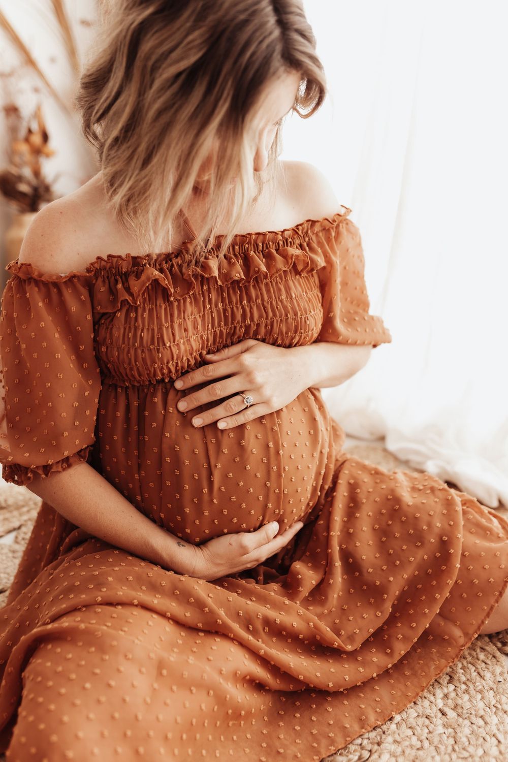 Side view of pregnant woman in rustic orange off-shoulder dress cradling baby bump while sitting on textured surface.