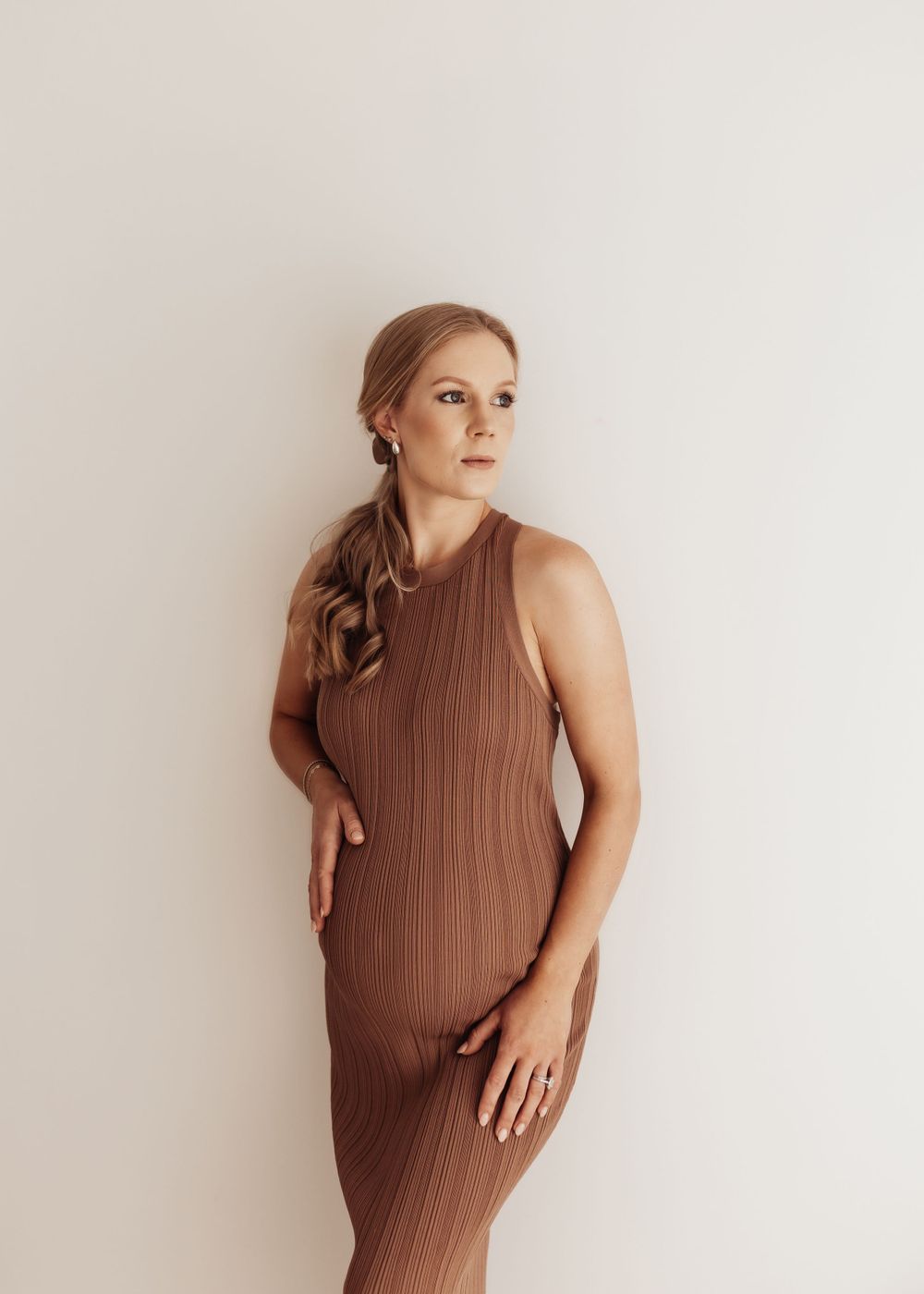 A pregnant woman in a sleeveless brown textured dress poses against a light beige wall for a maternity photo.