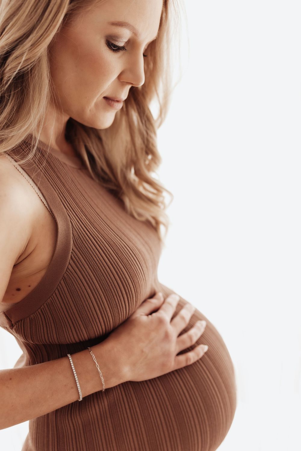 A person wearing a ribbed brown dress gently cradles their pregnant belly in a soft, minimalist maternity photo.