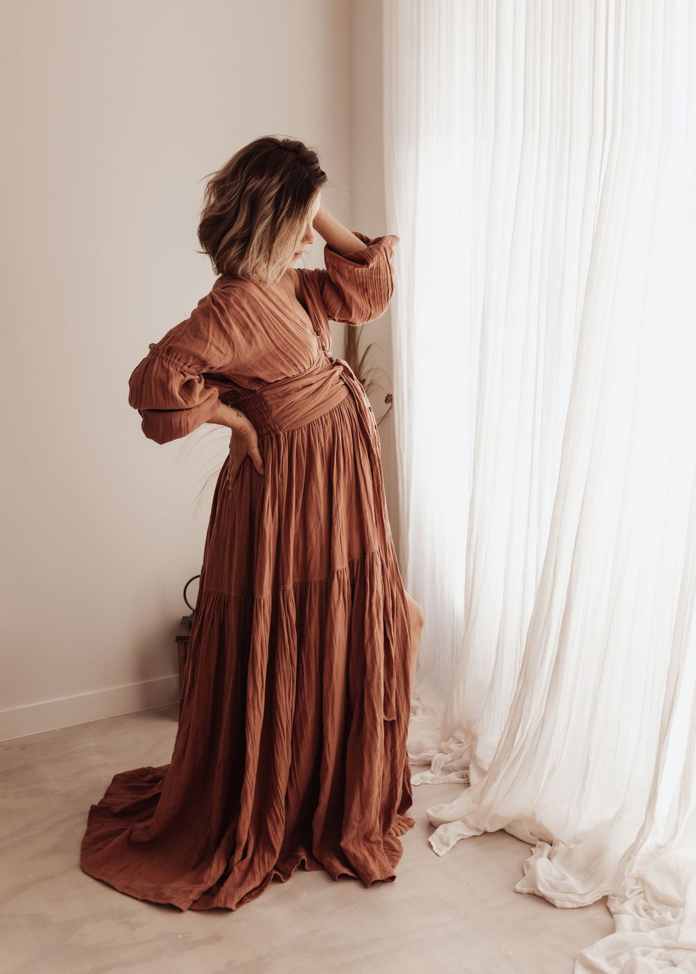 Figure in flowing rustic brown maxi dress with puff sleeves poses elegantly against sheer white curtains in sunlit room.