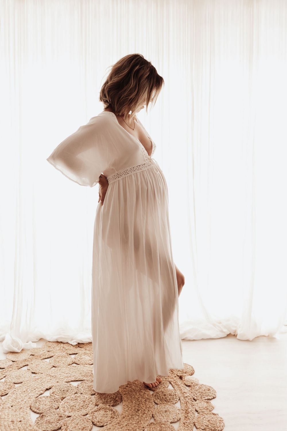 Silhouette of an expectant mother wearing a flowing white dress standing by sheer curtains in soft natural light.