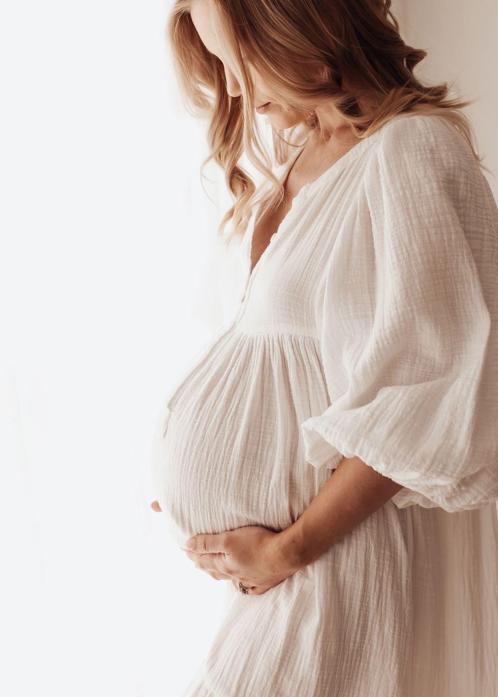 A woman in a flowing white cotton dress gently cradles her pregnant belly in soft natural light.