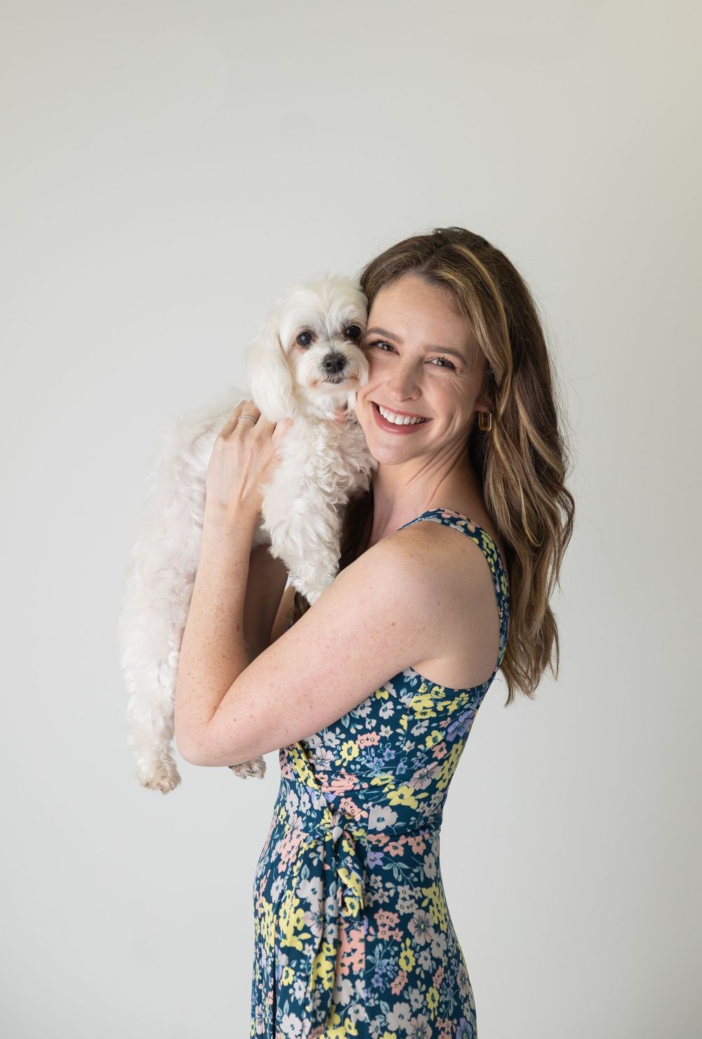 Someone wearing a floral dress holds a small white Maltese dog against a neutral background in a studio portrait style.