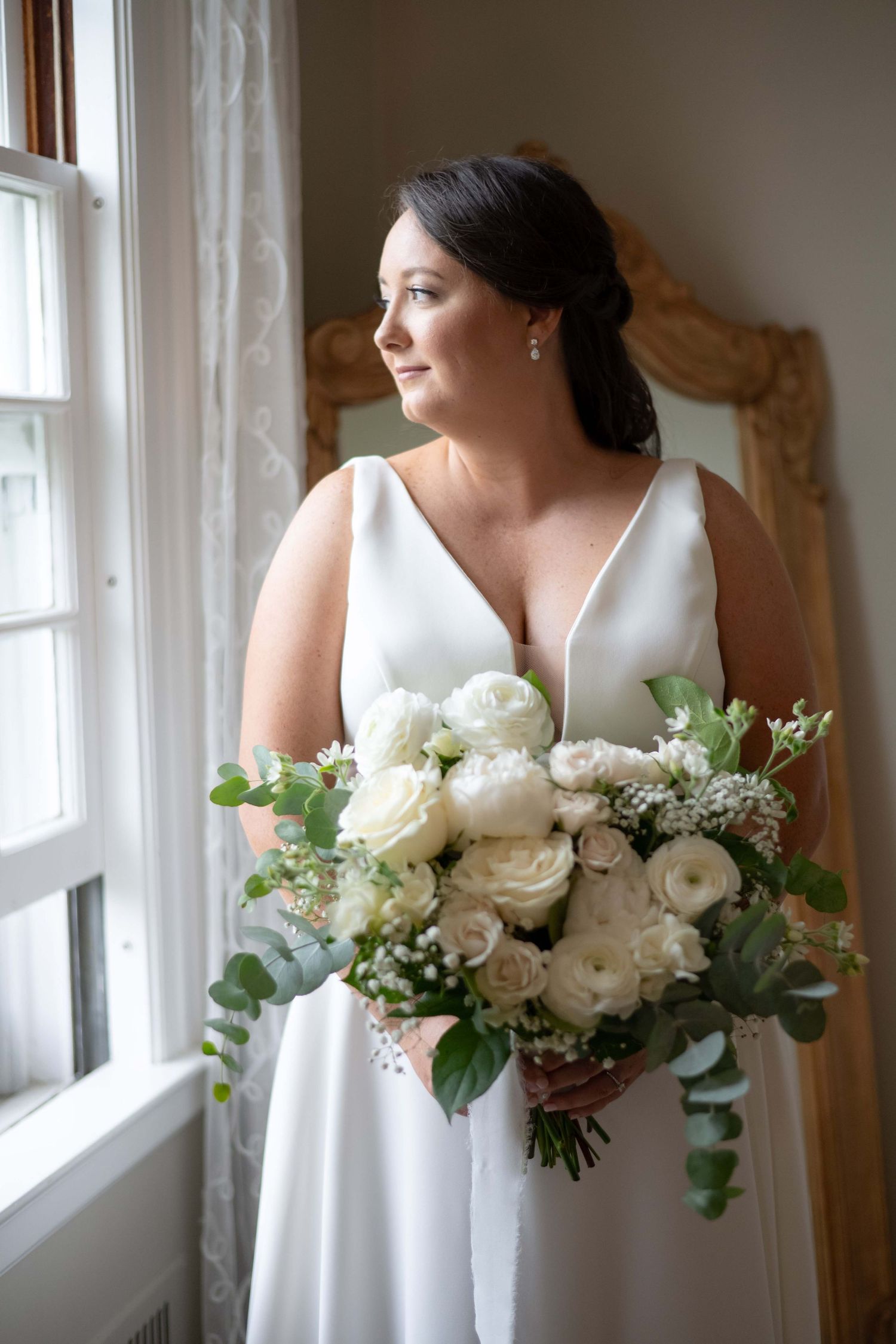 A bride in a classic white V-neck wedding dress stands by a window holding an elegant bouquet of white ranunculus and eucalyptus.