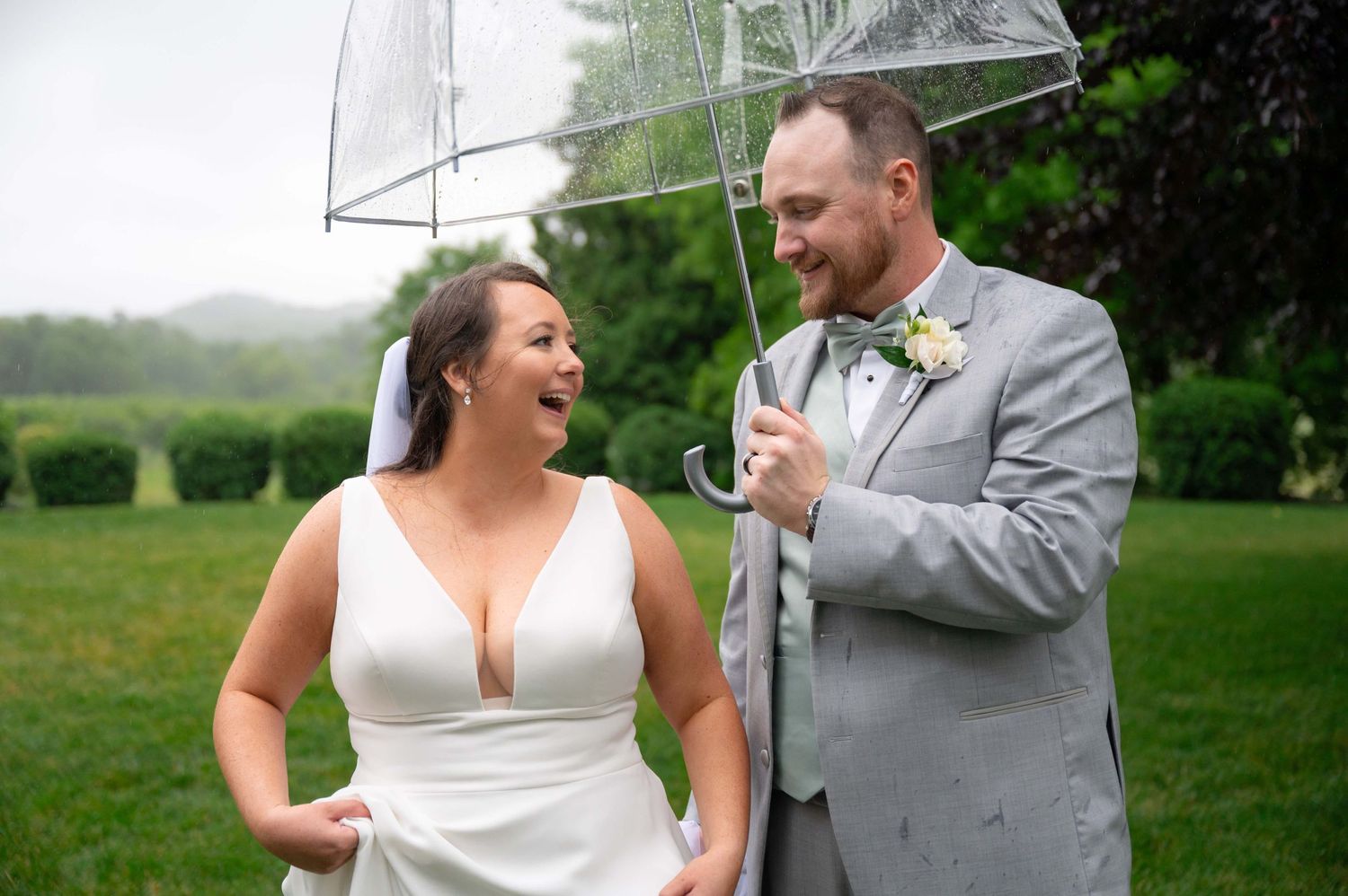 A happy couple laughs together under a clear umbrella during their rainy outdoor wedding celebration.
