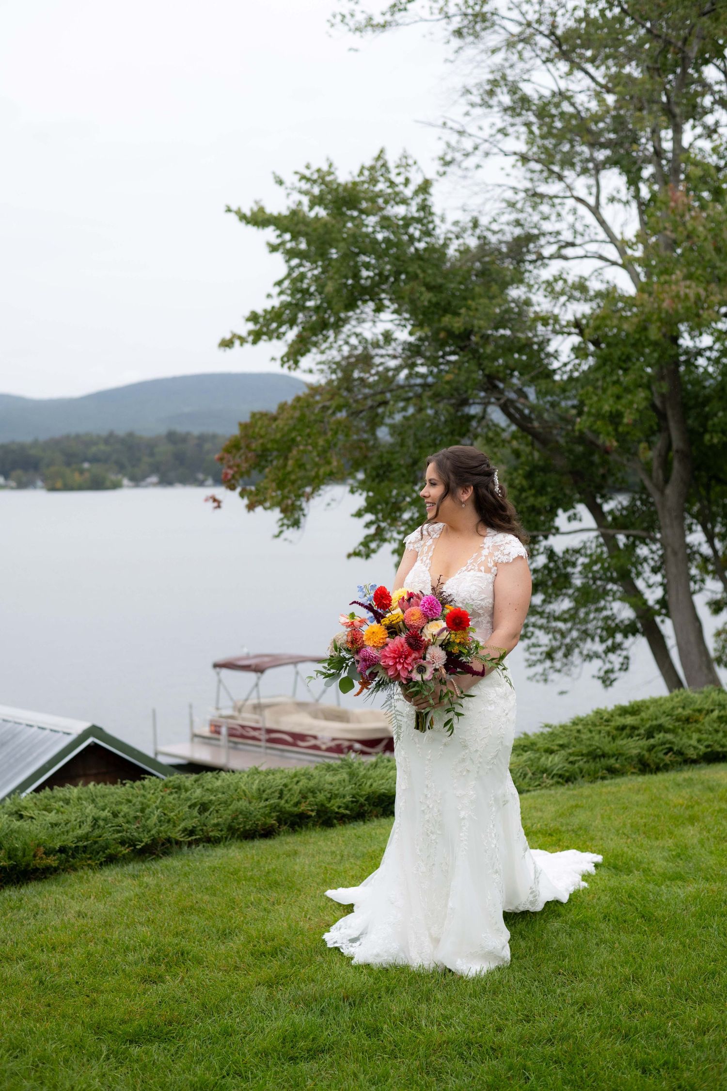 Bride in lace gown holds colorful bouquet while standing on lakefront lawn with mountain vista.