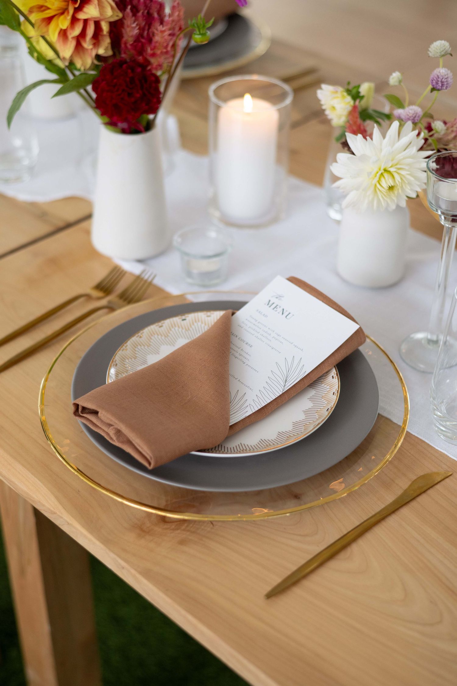 Elegant table setting with white ceramic vases, candle, and tan napkin on wooden table with gold-rimmed plates.