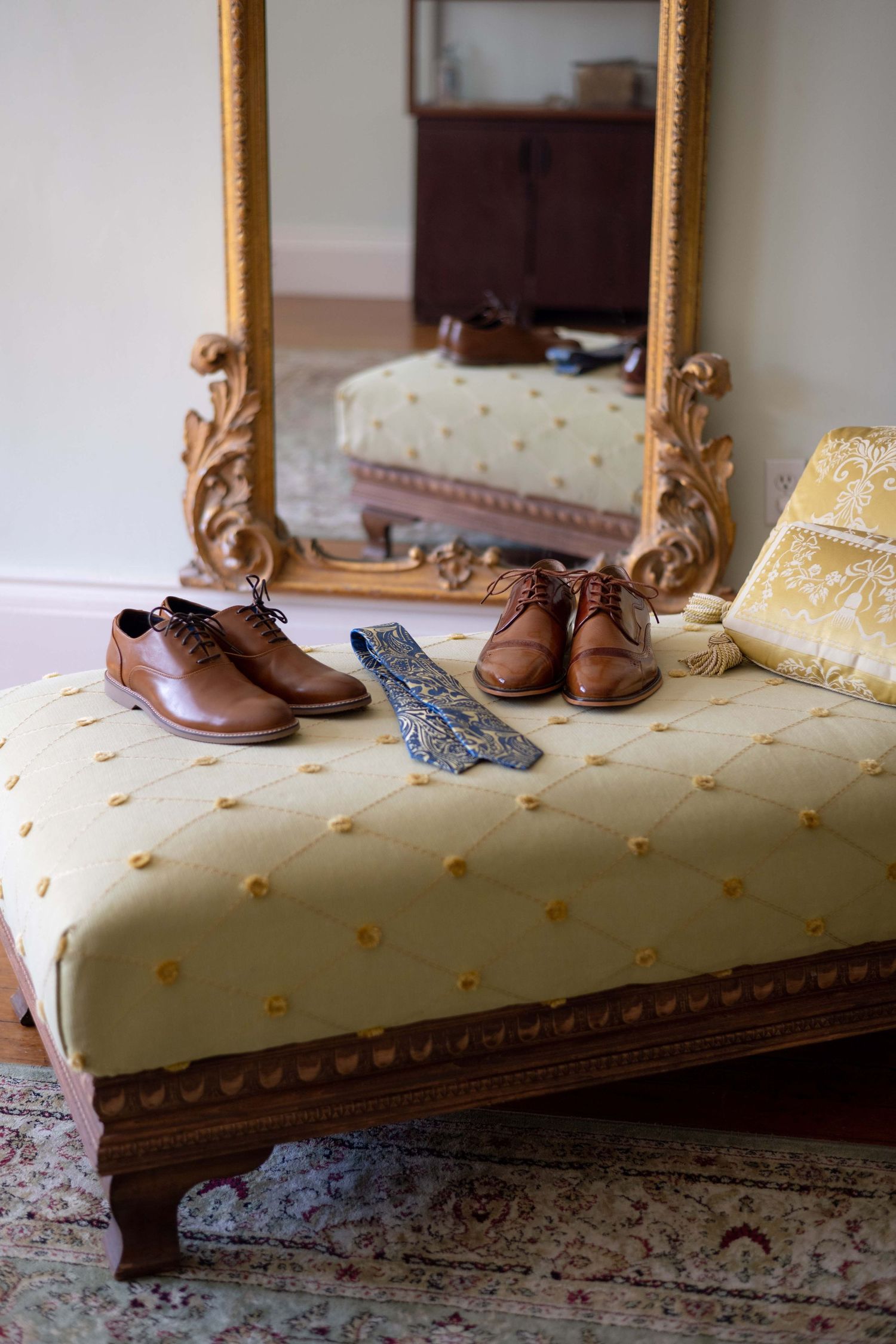 Brown leather dress shoes and blue tie arranged on a cream-colored upholstered ottoman with ornate wooden frame and mirror.
