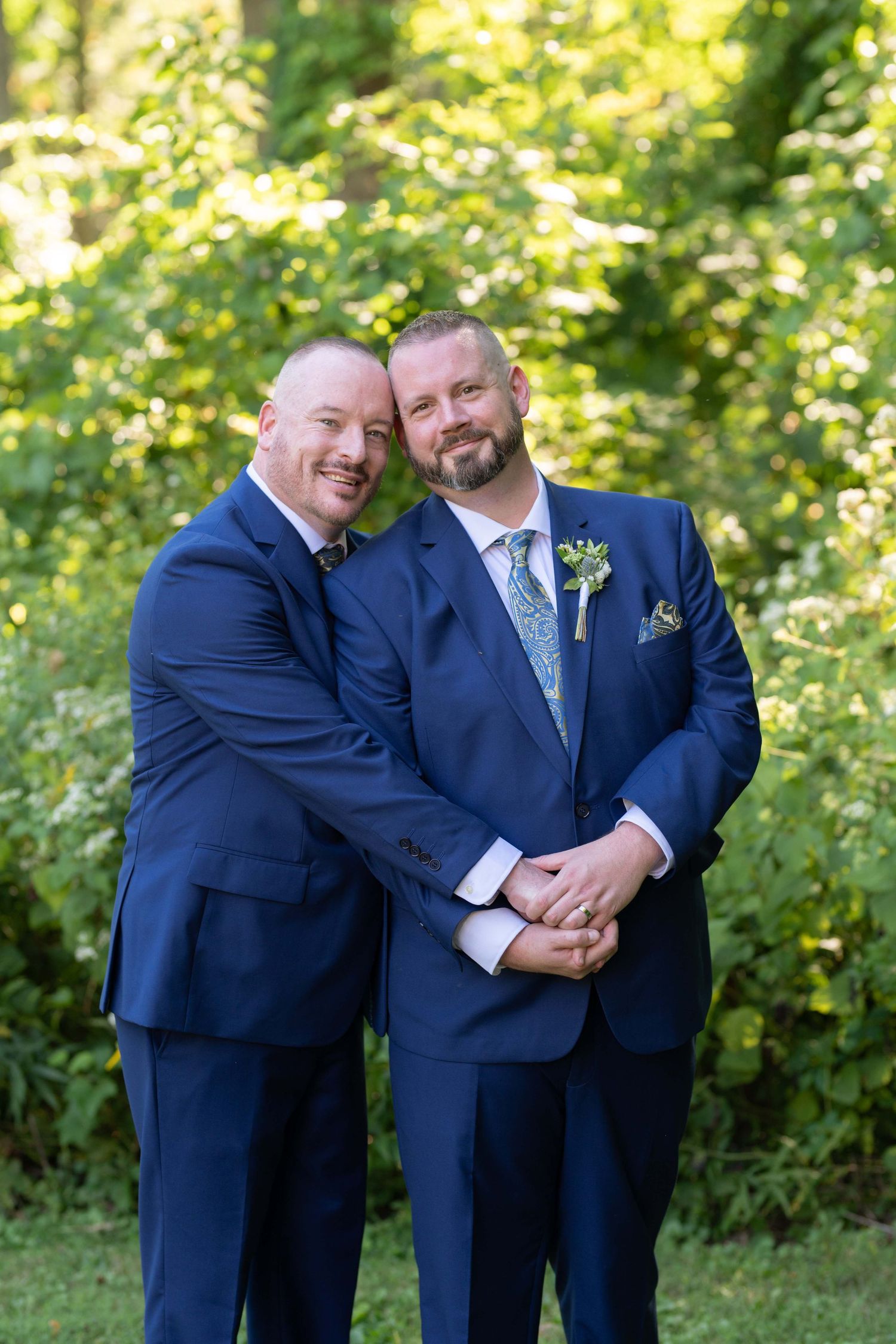 A couple in matching navy suits embrace outdoors on their wedding day with lush greenery in the background.