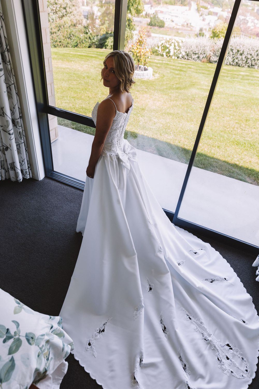 Bride in elegant white gown with long train standing by window.