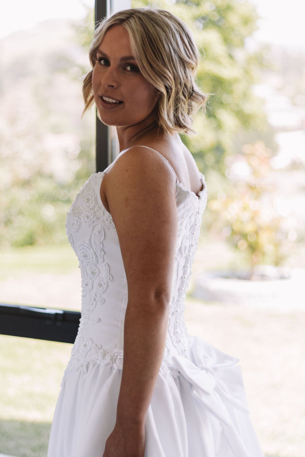A woman in a white wedding dress poses beside a window with natural lighting.
