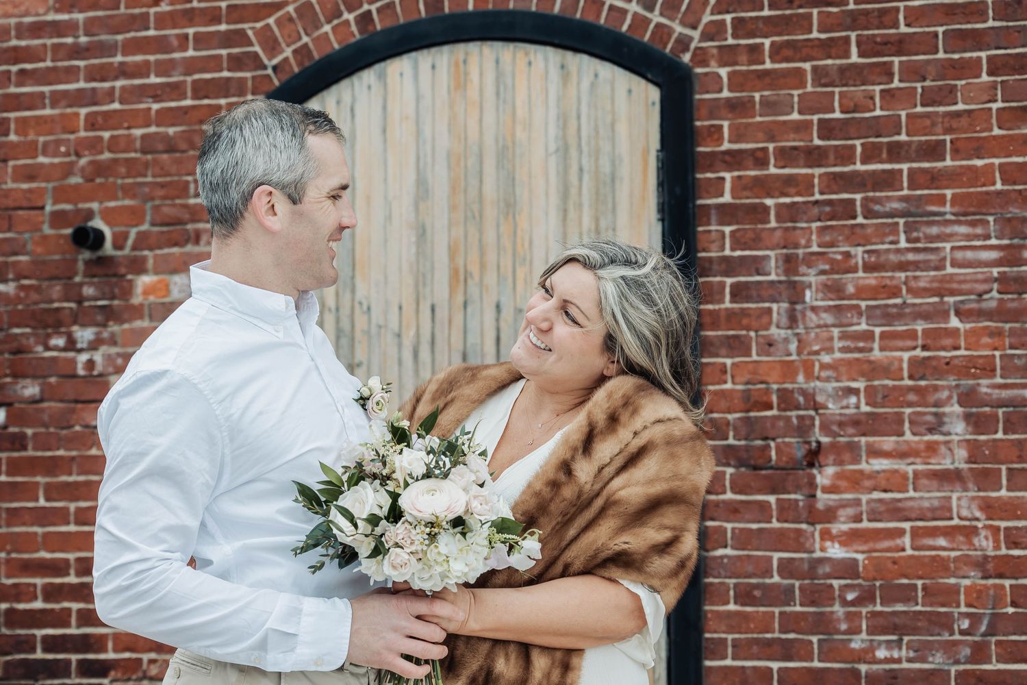 A romantic winter wedding portrait against a rustic brick wall with wooden door.